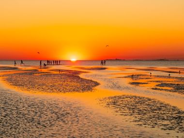 Goldener Sonnenuntergang über dem Strand Döse in Cuxhaven mit Silhouetten von Menschen auf nassem Sand und flachen Wasserbecken.