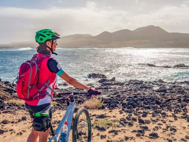Radfahrer mit grünem Helm und rosa Rucksack steht mit Fahrrad am vulkanischen Felsstrand auf Lanzarote mit Blick auf Meer und Berge.