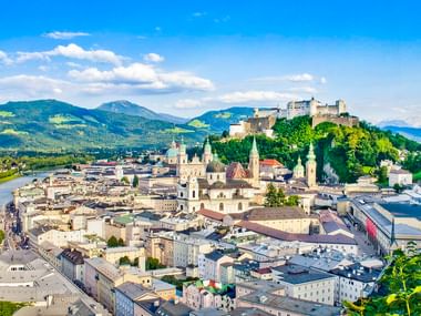 Panoramablick auf Salzburgs Altstadt mit Festung Hohensalzburg auf dem Hügel, der Salzach und Alpenbergen im Hintergrund.