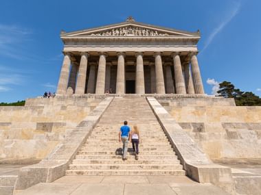 Zwei Radfahrer steigen die Treppe zur Walhalla bei Donaustauf hinauf. Der klassizistische Tempel mit dorischen Säulen steht auf einem Hügel.