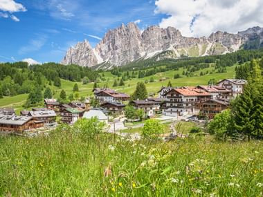 Alpendorf in Cortina d'Ampezzo mit traditionellen Chalets auf grünen Wiesen, umgeben von dramatischen Dolomitengipfeln unter blauem Himmel.