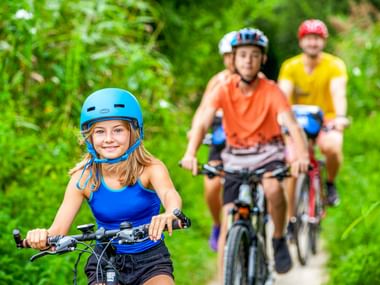 Junges Mädchen mit blauem Helm führt Familie auf Waldweg. Zwei Radfahrer folgen durch üppige grüne Vegetation in der Bretagne.