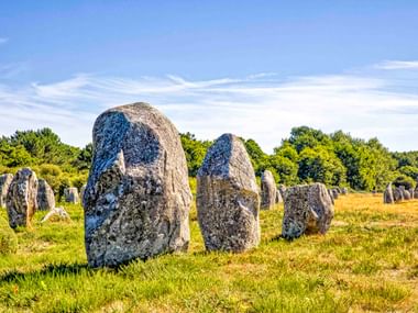 Antike Ménec Steinreihen auf einer Wiese in der Bretagne. Große verwitterte Megalithen stehen vor blauem Himmel mit Bäumen im Hintergrund.