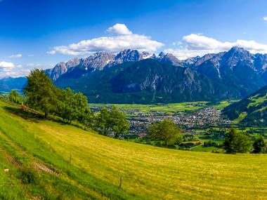 Grüne Wiesen und Bäume im Vordergrund mit der Stadt Lienz im Tal. Dramatische Dolomitengipfel unter blauem Himmel mit Wolken.