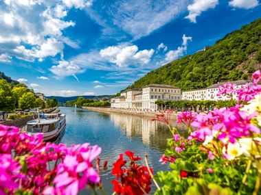 Bunte Blumen rahmen die Lahn in Bad Ems mit weißen historischen Gebäuden entlang der Promenade, grünen Hügeln und einem Boot auf dem Wasser.