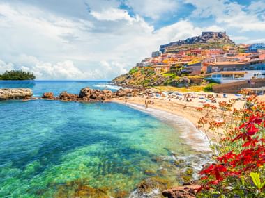 Strandbucht in Castelsardo mit klarem türkisfarbenem Wasser, roten Blumen im Vordergrund und Hangstadt mit historischer Burg auf der Spitze.