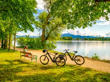 Person sitzt auf Bank am Chiemsee mit zwei Fahrrädern in der Nähe. Grüne Bäume rahmen den Blick auf den See mit Bergen in der Ferne.
