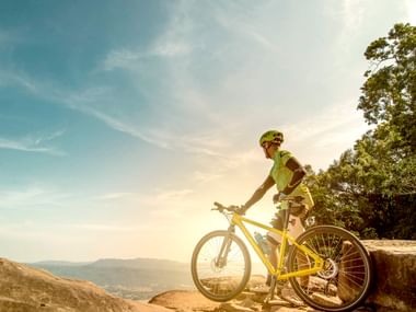 Radfahrer in grünem Trikot und Helm steht mit gelbem Mountainbike auf felsigem Aussichtspunkt mit Blick auf Tal und Berge unter blauem Himmel.
