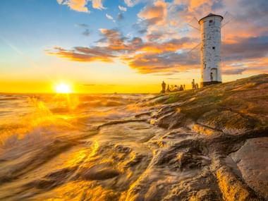 Weißer Mühlenbake Leuchtturm auf felsiger Küste in Swinemünde bei Sonnenuntergang. Goldene Wellen umspülen Felsen.
