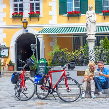 Zwei Radfahrer mit Tourenrädern rasten an einem Brunnen mit Statue vor einem gelben Gebäude mit roten Geranien in Oberdrauburg.