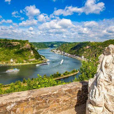 Steinstatue am Loreley Aussichtspunkt mit Blick auf den Rhein, der sich durch grüne Hügel schlängelt. Kreuzfahrtschiffe fahren auf dem Fluss.