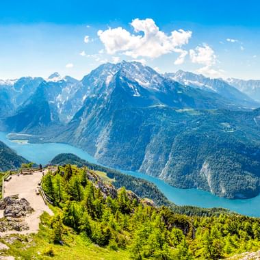 Luftaufnahme des türkisfarbenen Königssees zwischen bewaldeten Bergen. Eine Aussichtsplattform auf einem grünen Hang überblickt die alpine Landschaft.