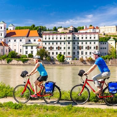 Zwei Radfahrer mit roten Rädern und blauen Taschen fahren am Inn in Passau entlang, mit historischen Gebäuden und einer Kirche am gegenüberliegenden Ufer.