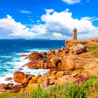 Der Leuchtturm Phare de Ploumanac'h steht auf rosa Granitfelsen an der bretonischen Küste. Blaue Meereswellen brechen sich an den Felsen.