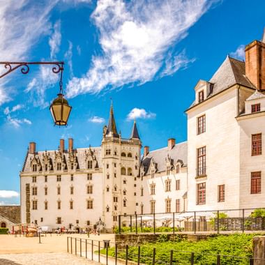 Weißes Château des Ducs de Bretagne in Nantes mit Türmen unter blauem Himmel. Innenhof mit Besuchern und historischer Laterne sichtbar.