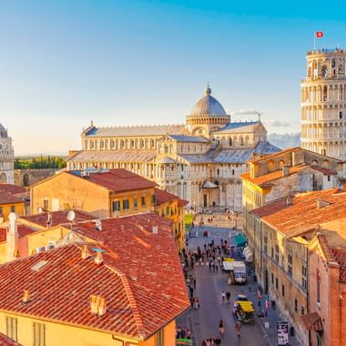 Luftaufnahme von Pisa mit Schiefem Turm, Dom und Baptisterium zwischen roten Ziegeldächern. Besucher gehen auf der Straße unter blauem Himmel.