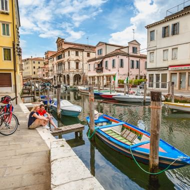 Zwei Radfahrer mit Rädern rasten an einem Kanal in Chioggia. Bunte Boote liegen an Holzpfählen. Historische Gebäude säumen das Ufer unter blauem Himmel.