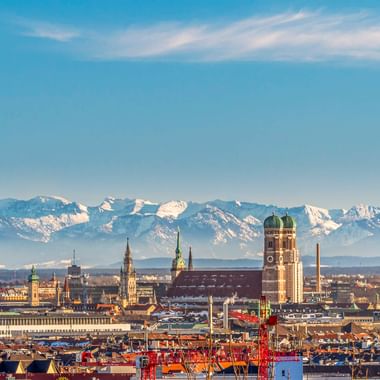 München Stadtansicht mit Frauenkirche-Türmen und historischen Gebäuden im Vordergrund, schneebedeckte Alpen im Hintergrund unter blauem Himmel.