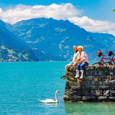 Zwei Radfahrer sitzen auf einer Steinmauer am Brienzersee mit einem weißen Schwan im türkisfarbenen Wasser. Berge und blauer Himmel im Hintergrund.
