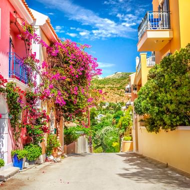 Schmale Straße auf Kefalonia mit rosa und gelben Häusern. Leuchtende Bougainvillea-Blüten hängen von Balkonen unter blauem Himmel.