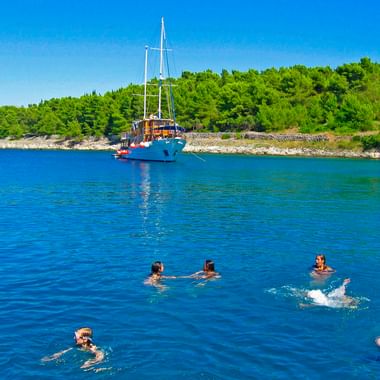 Sechs Personen schwimmen im klaren blauen Wasser nahe dem Segelschiff MS Mirabela, das in einer Bucht mit grüner bewaldeter Küste ankert.