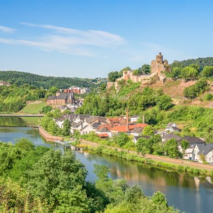 Panoramablick auf Saarburg mit Burgruine auf dem Hügel, Häusern entlang des Flusses, Weinbergen und grünen bewaldeten Hügeln unter blauem Himmel.