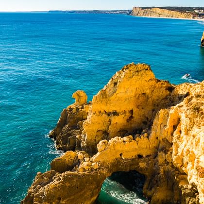 Goldene Sandsteinklippen ragen in das türkisfarbene Meer an der Algarve-Küste. Ein kleines Boot ist nahe den Felsformationen sichtbar.