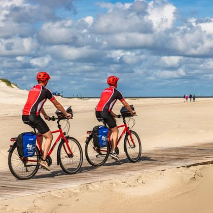 Zwei Radfahrer mit roten Helmen und Trikots fahren am breiten Sandstrand von St. Peter-Ording unter bewölktem Himmel. Sanddünen links sichtbar.