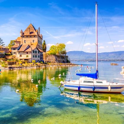 Blau-weißes Segelboot im klaren Hafen von Yvoire mit mittelalterlichen Steingebäuden und Kirche am Seeufer unter blauem Himmel.
