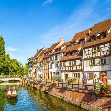 Bunte Fachwerkhäuser entlang eines Kanals in Colmar, Frankreich. Fußgänger spazieren auf der Uferpromenade, während ein kleines Boot auf dem Wasser treibt.