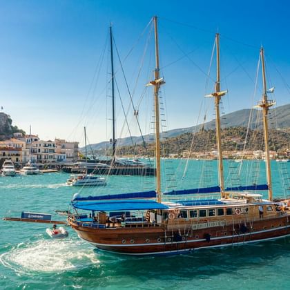 Hölzernes Segelschiff mit vier Masten in türkisfarbenem Hafenwasser. Griechische Küstenstadt mit weißen Gebäuden und Hügeln im Hintergrund.