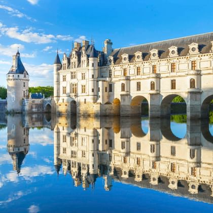 Schloss Chenonceau im Loiretal mit weißer Renaissance-Fassade und Bogenbrücke über ruhigem Wasser, perfekte Spiegelung unter blauem Himmel.
