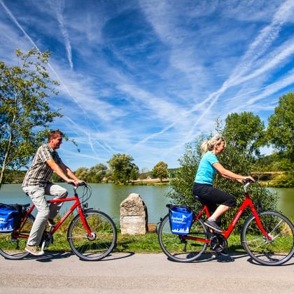 Zwei Radfahrer auf roten Rädern mit blauen Taschen fahren an einem See nahe der Altmühl-Quelle. Ein Steinmarker steht zwischen ihnen unter blauem Himmel.