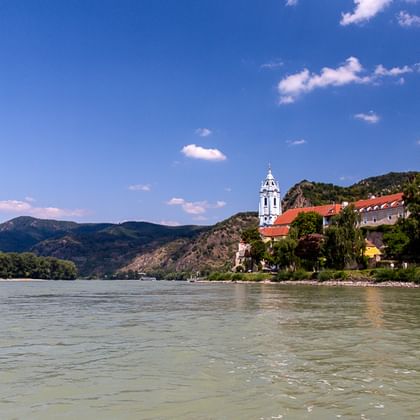 Blick auf die Donau mit dem blau-weißen Turm von Stift Dürnstein und Burgruine auf einem Hügel. Berge und blauer Himmel im Hintergrund.