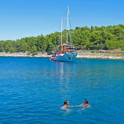 MS Mirabela, ein blaues Segelschiff mit zwei Masten, ankert in ruhigen kroatischen Gewässern. Zwei Schwimmer im Vordergrund, grüne Küste dahinter.