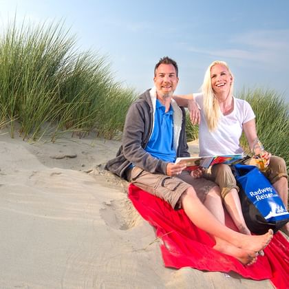 Lächelndes Paar sitzt auf roter Decke in Dünen in Zeeland mit Radkarte. Dünengras und Windräder im Hintergrund sichtbar.