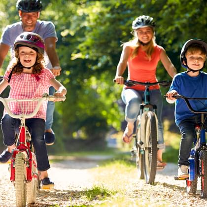Eltern und zwei Kinder fahren Rad auf einem von Bäumen gesäumten Weg. Alle tragen Helme. Mädchen in rosa Hemd auf rotem Rad, Junge in blau.
