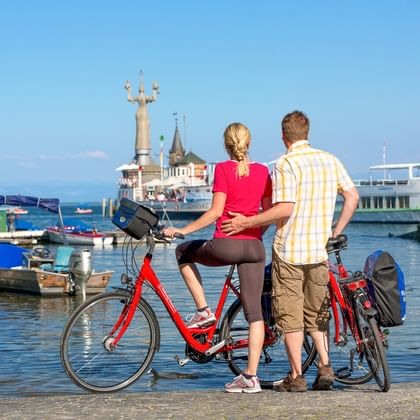 Paar mit Fahrrädern betrachtet den Hafen von Konstanz am Bodensee. Fähre und Imperia-Statue sichtbar, Boote im sonnigen Hafen.