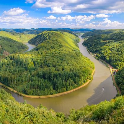 Luftaufnahme der Saarschleife bei Mettlach mit dramatischer Hufeisenbiegung der Saar, umgeben von dicht bewaldeten Hügeln unter blauem Himmel.