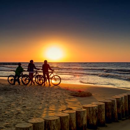 Drei Radfahrer mit Rädern stehen bei Sonnenuntergang am Sandstrand. Hölzerne Buhnen säumen das Ufer, Wellen rollen vom Meer herein.