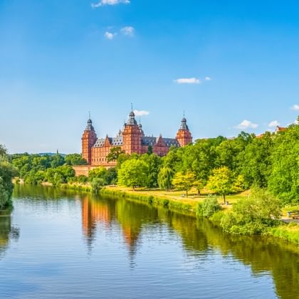 Schloss Johannisburg mit roten Sandsteintürmen spiegelt sich im Main, umgeben von grünen Bäumen unter blauem Himmel in Aschaffenburg.