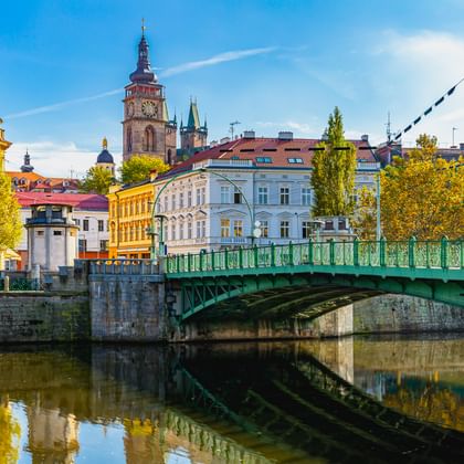 Grüne Zierbrücke über die Elbe mit bunten historischen Gebäuden und Kirchturm in einer tschechischen Stadt unter blauem Himmel.