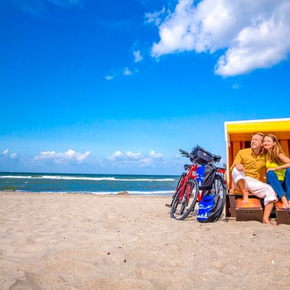 Paar entspannt in einem gelb-weißen Strandkorb mit zwei Fahrrädern daneben am Sandstrand in Wustrow unter blauem Himmel.