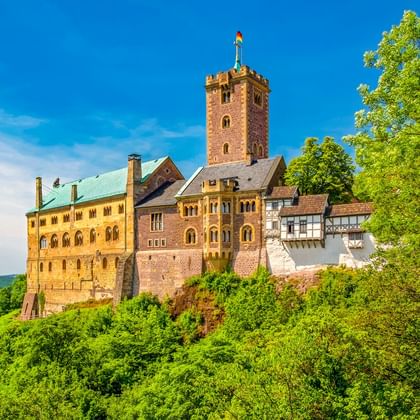 Die Wartburg in Eisenach thront auf einem bewaldeten Hügel mit rotem Turm, Steingebäuden und Fachwerkhäusern unter blauem Himmel.