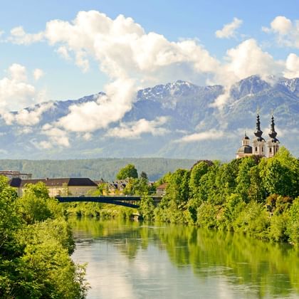 Blick auf Villach entlang der Drau mit grüner Vegetation, einer Kirche mit zwei Türmen und schneebedeckten Bergen unter bewölktem Himmel.