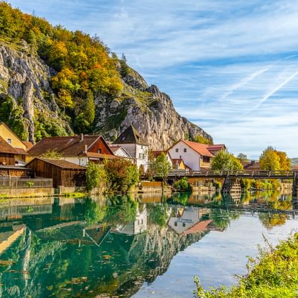 Dorf Essing am Altmühl-Fluss mit traditionellen Häusern, Holzbrücke und dramatischen Kalksteinfelsen in Herbstfarben.