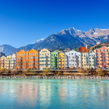 Reihe bunter historischer Gebäude entlang des türkisfarbenen Inn in Innsbruck mit schneebedeckten Berggipfeln im Hintergrund unter blauem Himmel.