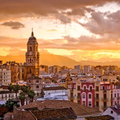 Sonnenuntergang über Málaga in Andalusien mit Kirchturm über weißen Gebäuden. Orangefarbener und goldener Himmel mit Bergen im Hintergrund.
