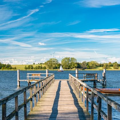 Holzsteg, der in die Schlei führt, mit festgemachten Booten. Blaues Wasser, grünes Ufer mit Bäumen und Windrädern unter hellem Himmel.