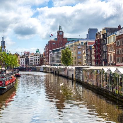 Amsterdamer Gracht gesäumt von bunten historischen Gebäuden unter bewölktem Himmel. Eine Brücke überquert das Wasser, Boote liegen am Ufer.